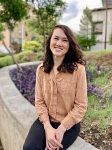 A photograph of Miranda Nadeau, PhD AAPI therapist. She is smiling and making eye contact with the viewer. She wears a burnt orange striped shirt with black pants and gold earrings. Behind her, out of focus, is a lawn with grass, purple heart, and trees. A cloudy sky is visible above.